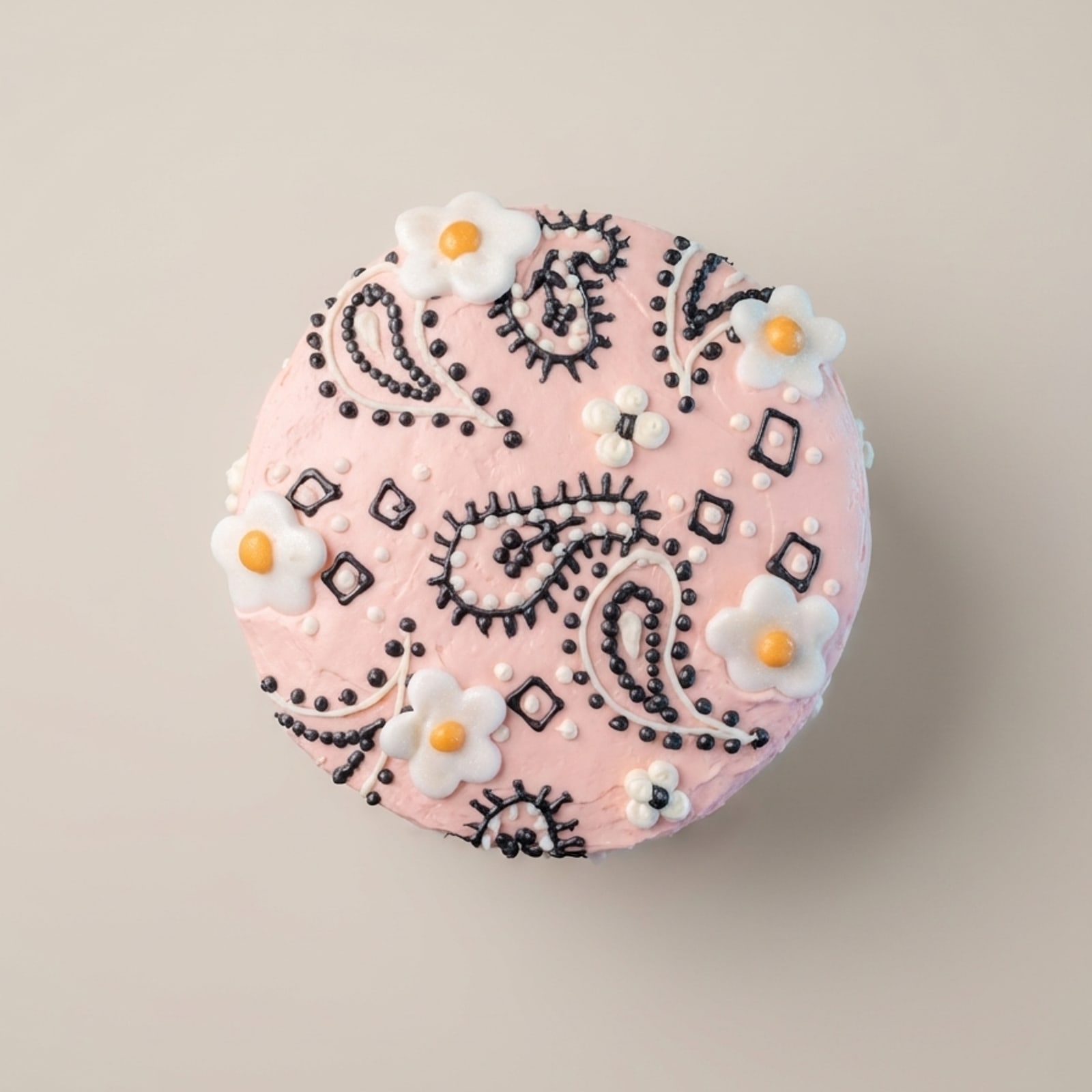 Top view of a round cake with pastel pink frosting, dotted black-and-white paisley, and white blossom accents.